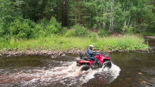 Drone shot of man in riding gear driving red quad bike through river in forest