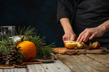 Chef cuts with knife apple on wooden chopped board for preparing aromatic muled wine with fragrant species on christmas composition background. Backstage of making hot winter drink. Traditional recipe