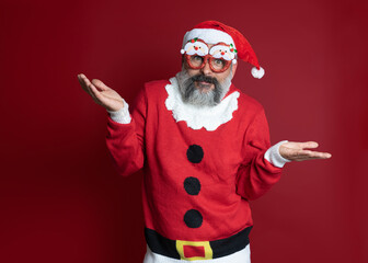 Fototapeta na wymiar Man in a Christmas sweater with party glasses spreading hands in surprise on red background