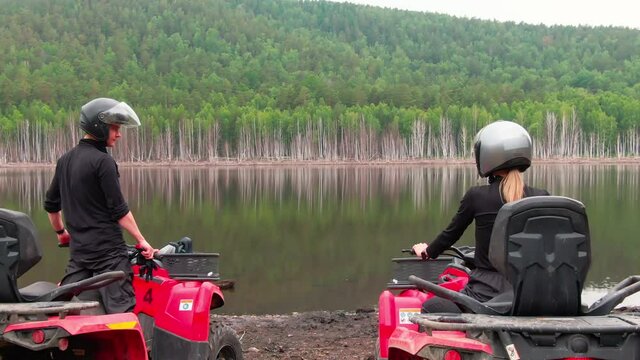 Aerial shot of couple in helmets sitting on red quad bikes by the lake in picturesque area
