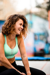 cheerful woman with curly hair in bright sports top sitting on trampoline