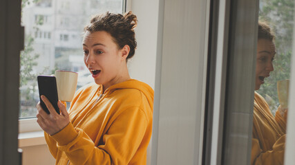 Excited surprised young brunette woman in yellow hoodie with cup of coffee holding mobile phone and...
