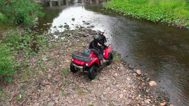 Aerial shot of happy man and woman in riding gear sitting on red quad bike by the river in forest and chatting