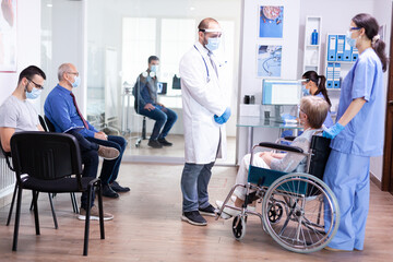 Young doctor wearing face mask against coronavirus talking with disabled woman in wheelchair during examination against covid-19. Young man waiting for medicconsultation. Health care system.