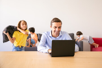 Cheerful dad chatting via laptop and kids playing with pillows near him. Caucasian father working at home during school vacations. Happy children on background. Family and digital technology concept