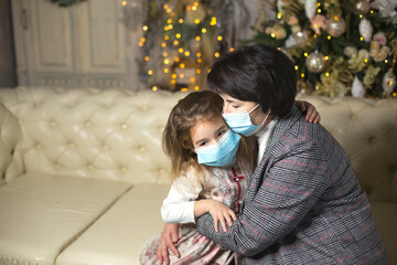 Grandmother and granddaughter on the sofa in the living room with Christmas decor hugging in medical masks on their faces. A family holiday during the outbreak of coronavirus and disease. New Year