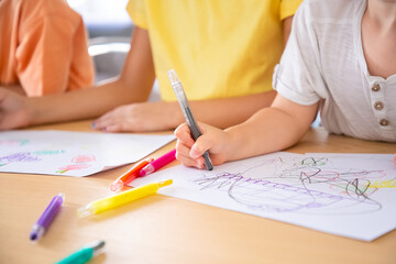 Cropped view of children painting on paper with pens. Three unrecognizable kids sitting at table...