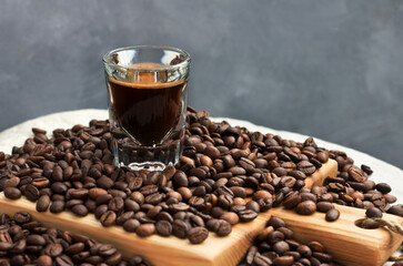 A short transparent glass of coffee situated amon scattered,  roasted and calibrated coffee beans on a wooden cutting board, napkin, grey background, close up