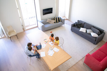 Top view of father and children sitting at table together. Two kids painting doodles with colorful pens. Middle-aged dad using laptop and holding little son. Childhood, interior and family concept