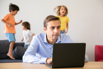 Serious Caucasian dad working via laptop and kids jumping on background. Concentrated father using computer and kids playing on sofa. Selective focus. Childhood and digital technology concept