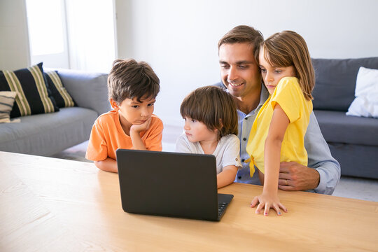 Happy Dad Sitting At Table, Embracing Kids And Using Laptop. Caucasian Middle-aged Father Browsing In Internet With Lovely Children. Childhood, Fatherhood And Digital Technology Concept