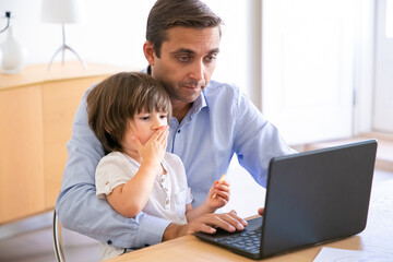 Focused father using laptop and holding son on knees. Caucasian middle-aged dad sitting at table with cute little boy and working on computer. Childhood, freelance and fatherhood concept