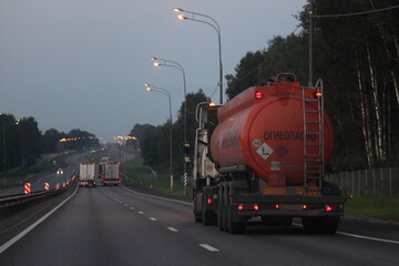 Modern orange semi truck fuel tanker with 30 1202 dangerous class sign and Russian inscription INFLAMMABLE drive on suburban road at summer evening in perspective, rear side view ADR hazardous cargo