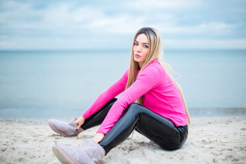 Fototapeta na wymiar blonde caucasian female with blue eyes in patent leather trousers and crimson pullover is sitting on the sand at the beach, dramatic sky and sea at the background
