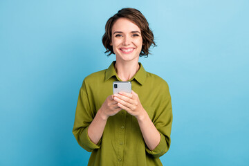 Photo of pretty lovely lady short hairstyle arms hold telephone cheerful shiny white smile texting email wear green shirt isolated blue color background