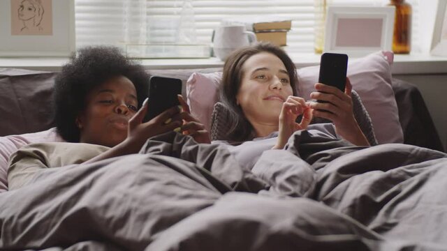 Young African American and Caucasian girlfriends lying together on bed, scrolling social media on their smartphones, talking and smiling in the morning at home