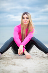 Fototapeta na wymiar blonde caucasian female with blue eyes in patent leather trousers and crimson pullover is sitting on the sand at the beach, dramatic sky and sea at the background