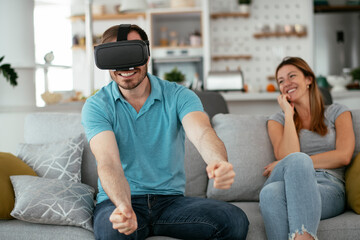 Fototapeta premium Smiling young man using VR headset at home on couch. Woman and her husband enjoying virtual reality in apartment.