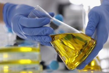 Hands in blue gloves hold flask with clear golden liquid in laboratory. Oil research and their...