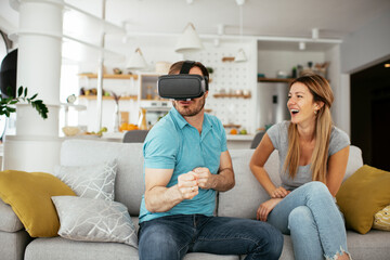 Fototapeta premium Smiling young man using VR headset at home on couch. Woman and her husband enjoying virtual reality in apartment.