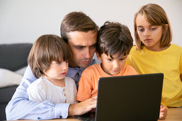 Fototapeta na wymiar Concentrated father and serious kids looking at screen. Caucasian middle-aged dad typing on laptop screen and children watching his work. Fatherhood, childhood and digital technology concept