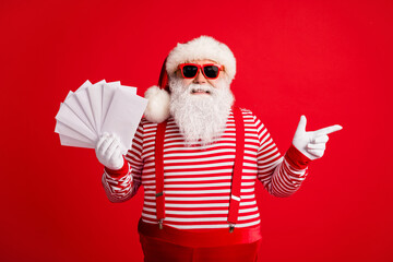 Fototapeta na wymiar Photo of grandfather grey beard direct finger empty space hold fan envelopes wear santa claus x-mas costume suspenders sunglass striped shirt cap gloves isolated red color background