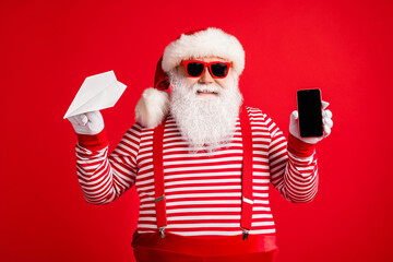 Photo of old man grey beard hold origami plane telephone screen empty space wear santa claus x-mas costume suspenders sunglass striped shirt cap isolated red color background