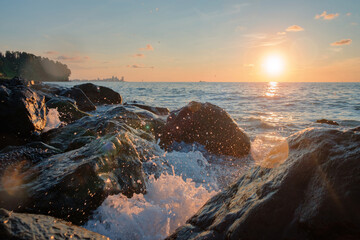 Sea waves crushing over the big rocks with beautiful sunset sky and city line on the background