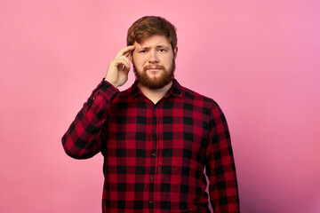 Portrait of a bearded man. The red-haired guy in a plaid shirt is thinking, holds his hand to his head. on isolated pink background