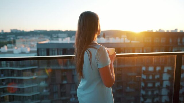 Woman with a cup of coffee standing on the balcony and admire the sunset