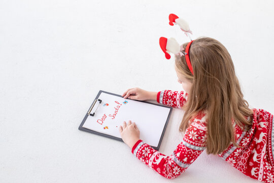 Child writing letter to Santa Claus at Christmas on white background. Top view. notepad for wishes, gift list.Girl in red dress lying and writing christmas letter.celebration concept. - Powered by Adobe