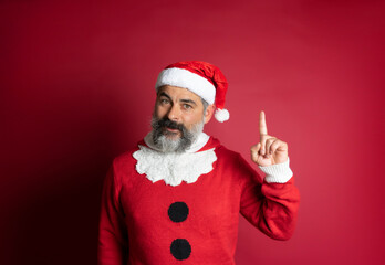 Fototapeta na wymiar Bearded man wearing christmas hat pointing finger up looking at camera on red background