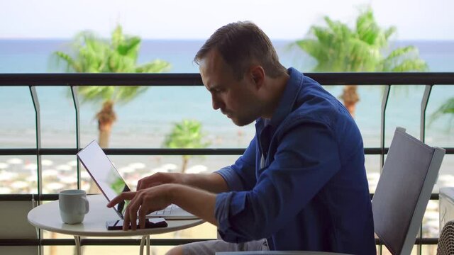 Businessman sit on balcony work on laptop at tropical beach resort. Young professional male freelancer answering cellphone to discuss business project on screen with colleugue from remote home office