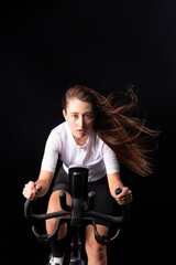 Fototapeta na wymiar Young female indoor cyclist grips the handlebars as she rides against the wind during stationary bicycle fitness workout. Front view, dark background.