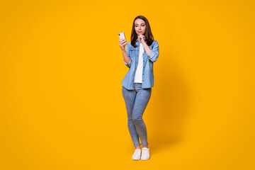 Full length photo of attractive cheerful lady good mood chatting telephone friends think over creative blog post theme wear casual denim shirt sneakers isolated vivid yellow color background