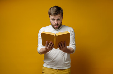 Attractive young man enthusiastically reads a book, standing against a yellow background. Education...