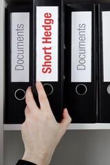 Short Hedge. File Folder is taking by a hand from office shelf. Red Text is on the label of the...