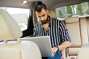 Successful arab man wear in striped shirt and sunglasses pose inside white suv car with laptop in hands.