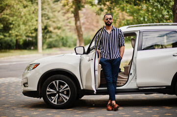 Successful arab man wear in striped shirt and sunglasses pose near his white suv car. Stylish...