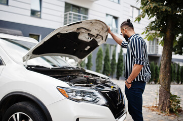 Successful arab man wear in striped shirt and sunglasses pose near his white suv car, check engine with open hood.