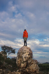 man hiker with orange jacket standing on top of stone and enjoying amazing view on bright blue sky. Travel and active lifestyle concept.