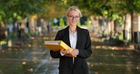 A nice girl in glasses, leaning on her right hand, holds a document folder and smiles cutely, looking at the camera.