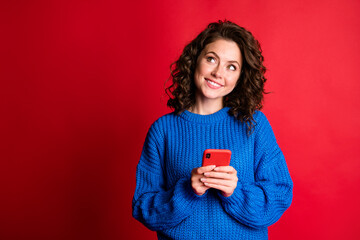 Photo of interested charming pretty lady wavy hairdo hold telephone look up empty space toothy beaming smiling wear blue knitted sweater pullover isolated red color background