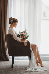A blonde bride with a high hairstyle is sitting on a chair opposite a large window with a bouquet. Long, slender legs are visible.