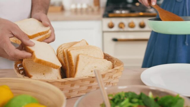 Tracking right of unrecognizable man putting slices of bread into breadbasket while unrecognizable woman putting friend egg with bacon on plate and then taking small tomato in hand
