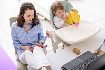 smiling brunette woman with graphic tablet in hands sitting at laptop, child with toy sitting next to her, Working from home