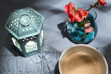 Selective focus flat lay view of bowl of coffee, lighted lantern, and small vase of flowers on a background of pale blue fringed shawl