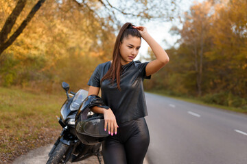 Fototapeta na wymiar Female motorcyclist stands near motorcycle on roadside and holding helmet