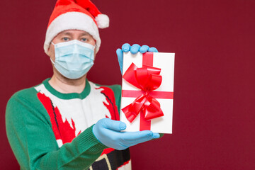Handsome male wears Santa hat medical mask gloves holds gift box showing on camera red studio background.