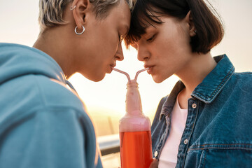 Cropped shot of happy young lesbian couple drinking from one glass bottle with the straw, Two girls enjoying cold beverage on a summer day outdoors